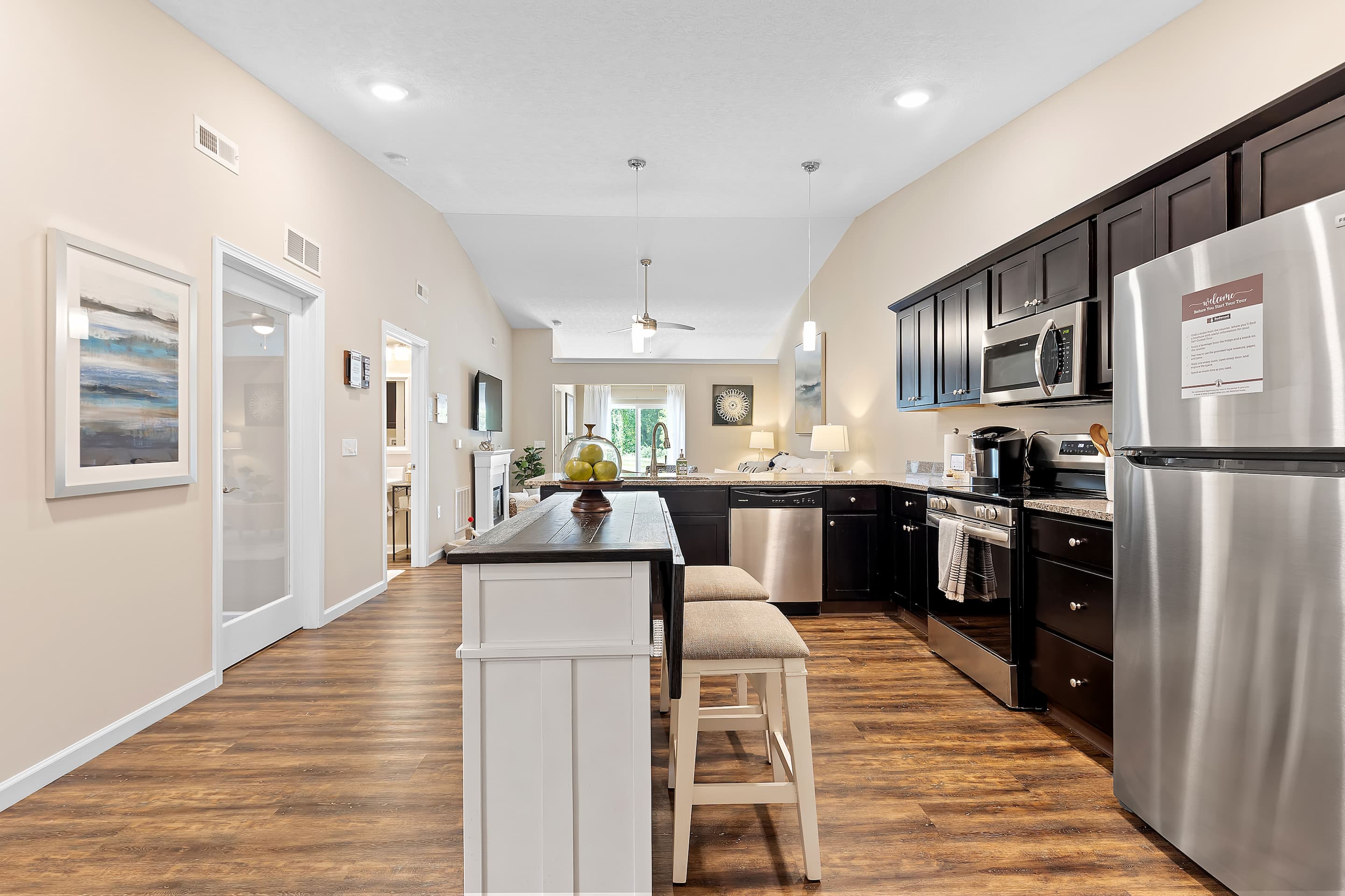 Modern kitchen with dark cabinets, stainless steel appliances, an island with two stools, wood flooring, and an open view into a living area with a large window.