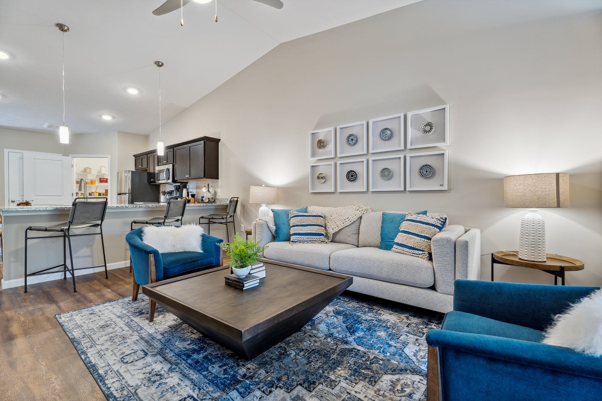 A modern living room with a gray sofa, two blue chairs, a patterned rug, and a wooden coffee table. The kitchen features black cabinetry, a counter with bar stools, and pendant lights.