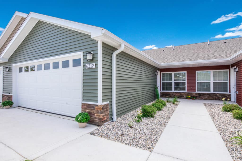 A single-story house with a green garage door, stone accents, and a pathway leading to the entrance, located at house number 6312. The exterior features neatly landscaped rocks and plants.