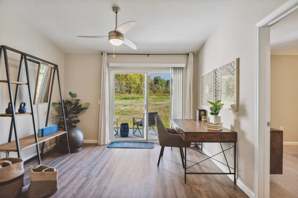 A home office with a wooden desk, chair, and a plant. A sliding glass door leads to an outdoor view. Shelving and a ceiling fan are in the room, with wooden floors and light walls.