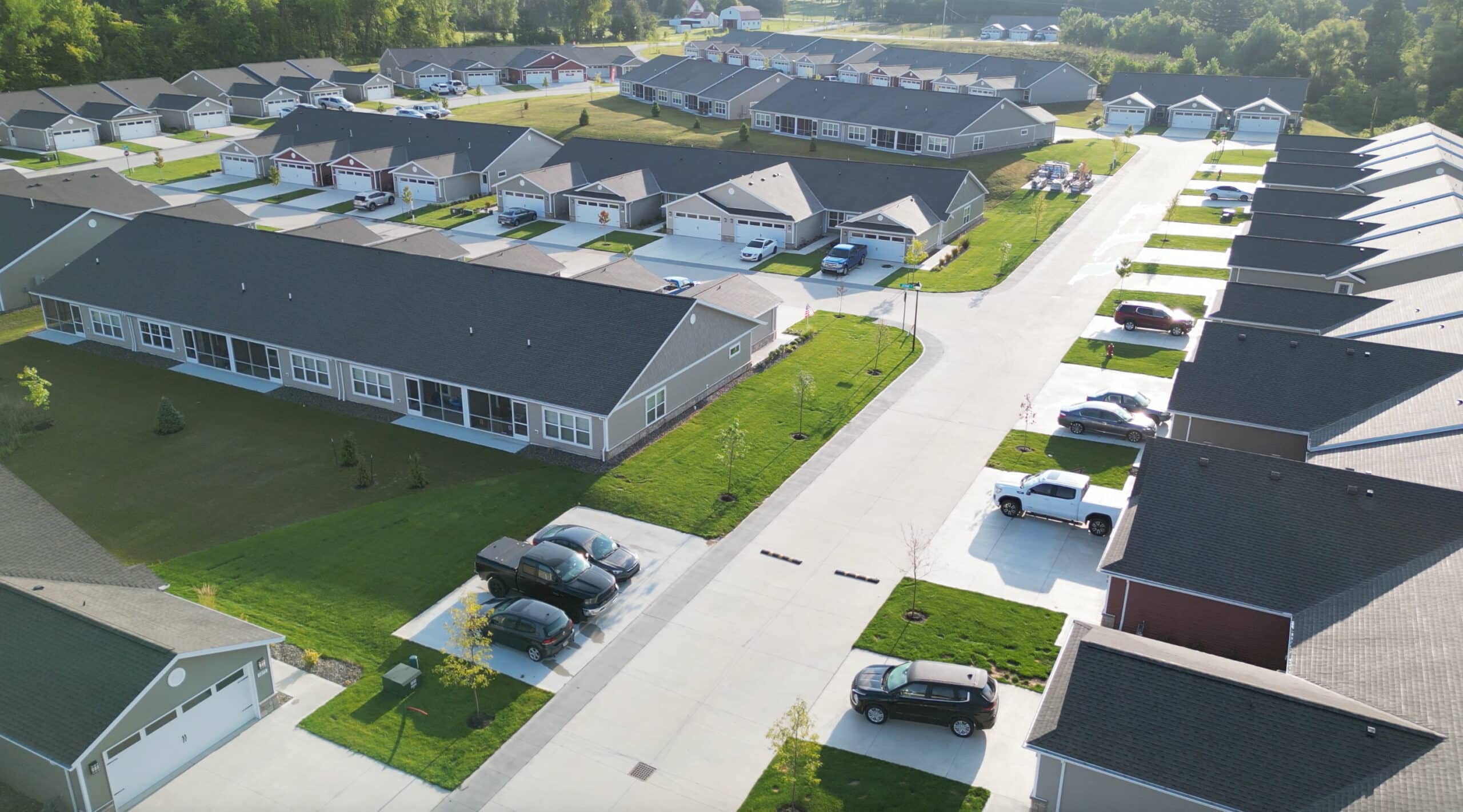 Aerial view of a residential neighborhood featuring multiple single-story houses with driveways, parked cars, spacious lawns, and a main street running through the area.