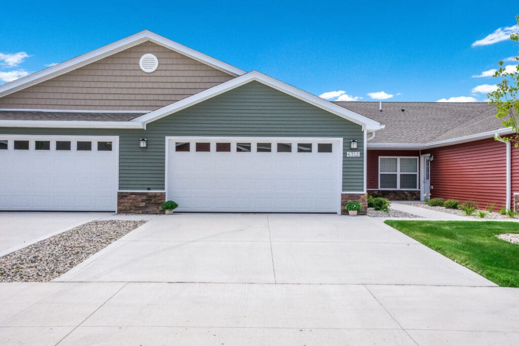 A residential building with green and red exterior siding, two white garage doors, driveway, and small landscaped areas with plants and rocks under a clear blue sky.