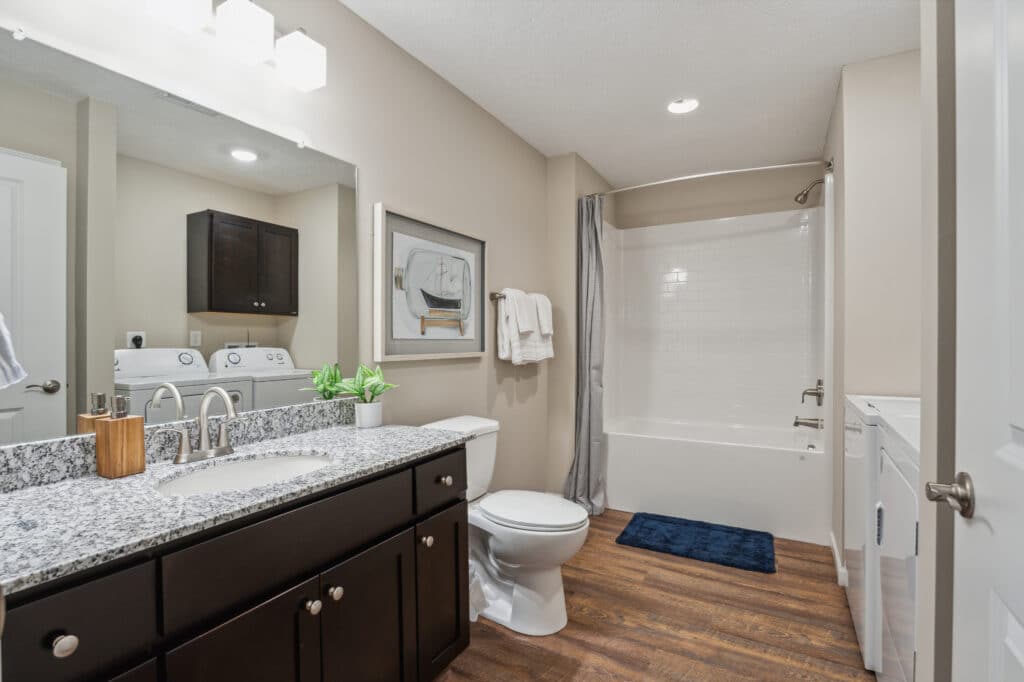 A modern bathroom featuring a granite-top vanity, toilet, washing machine, dryer, and a shower-tub combo with a white tiled wall and gray curtain, all set on a wood-patterned floor.