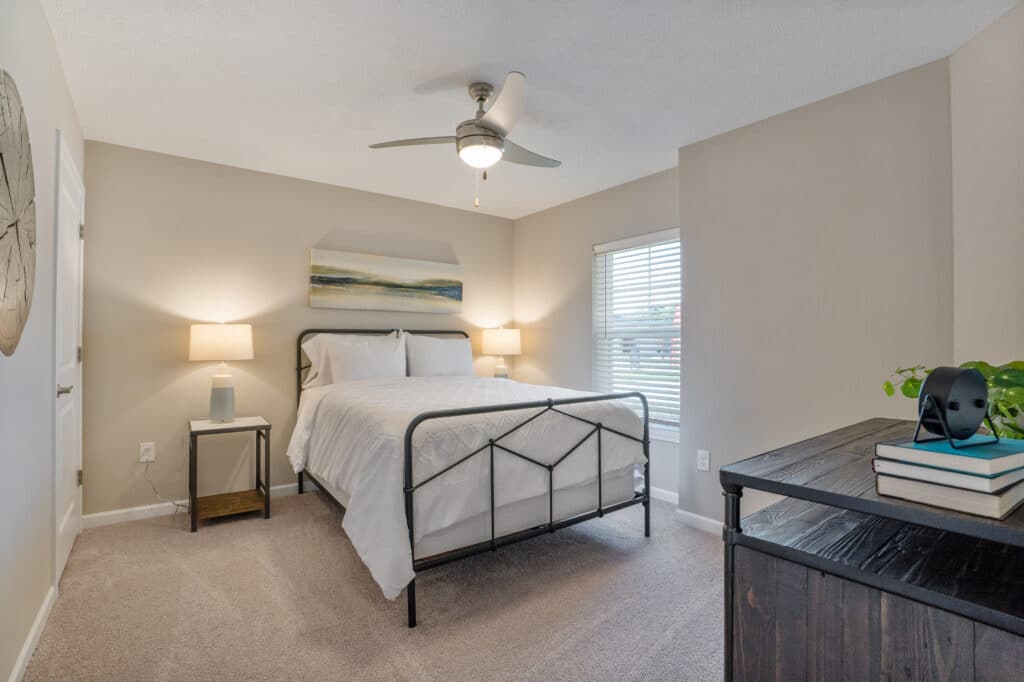 A minimalist bedroom with a bed, two bedside tables with lamps, a ceiling fan, a dresser topped with books, a potted plant, and a window with blinds. Beige walls and carpeted floor.