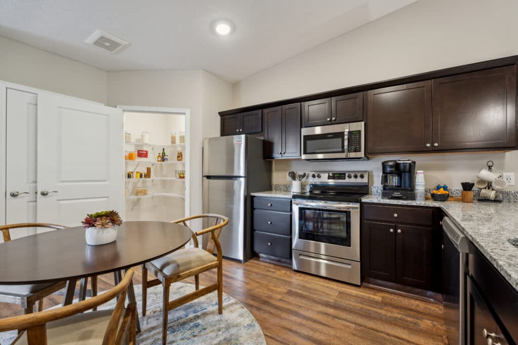 A modern kitchen with stainless steel appliances, dark wood cabinets, granite countertops, and a round dining table with chairs. A pantry with open doors is visible in the background.