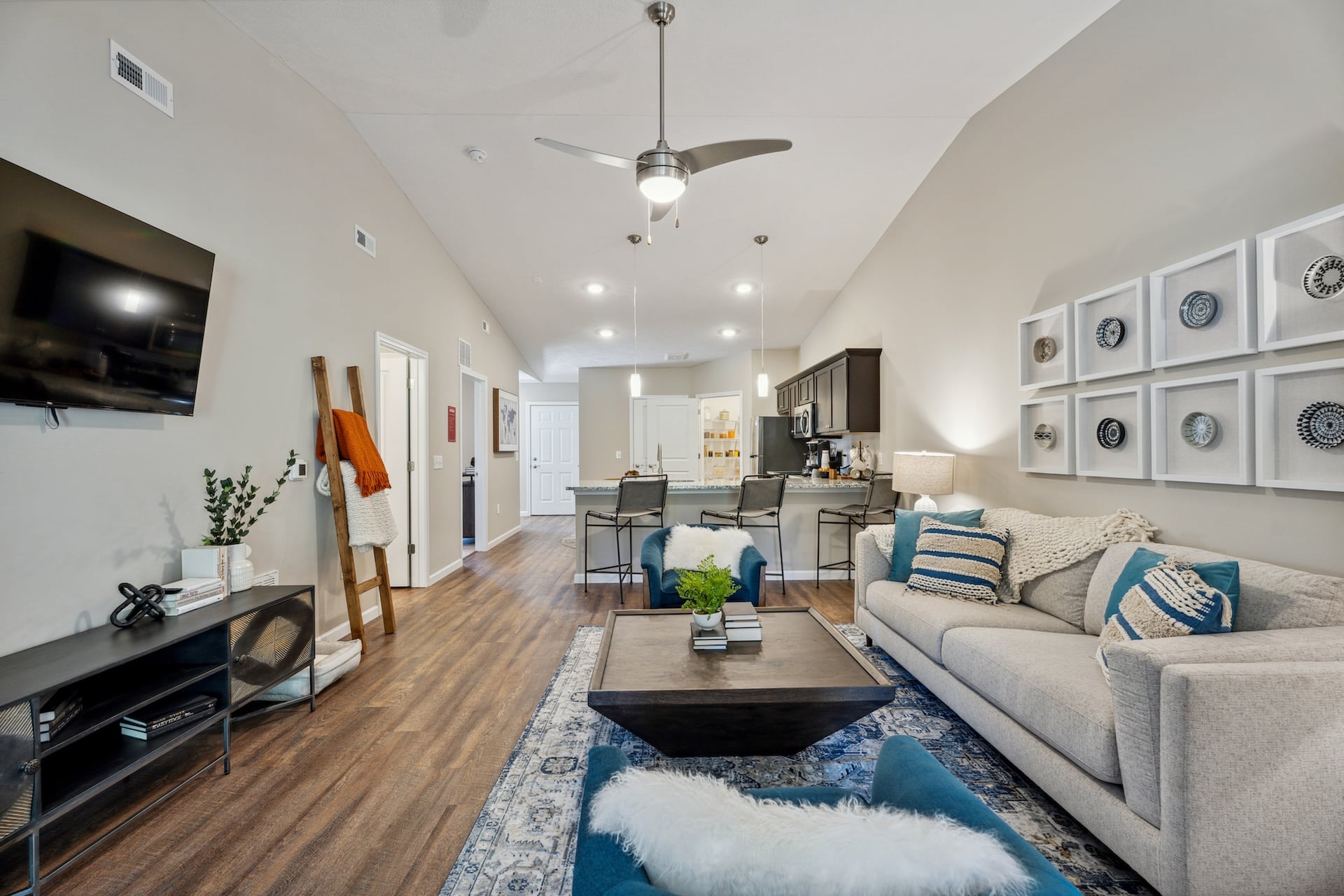 A modern living room with light gray walls, wood flooring, a beige sofa, blue and white decor, a wall-mounted TV, and an open kitchen in the background with bar stools at a counter.