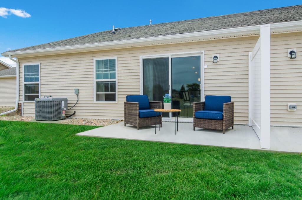 A patio with two wicker chairs with blue cushions and a small table sits on a concrete slab outside a beige house with a sliding glass door.