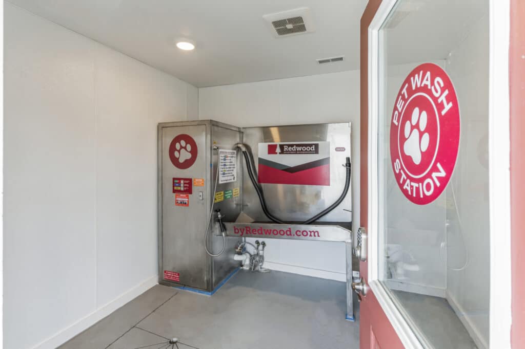 A small room with a "Pet Wash Station" featuring a large metallic washing unit, hoses, and a red sign with a paw print logo. The door and wall also display "Pet Wash Station" signs.