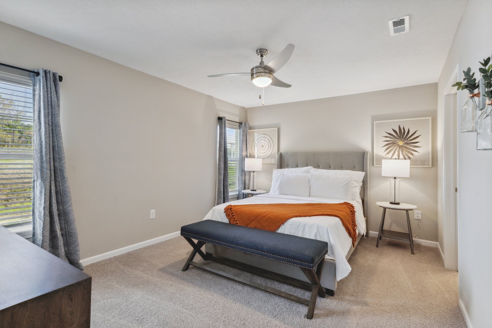 A bedroom with a bed, white linens, an orange throw, two bedside tables with lamps, a ceiling fan, two windows with blinds, and a cushioned bench at the foot of the bed.