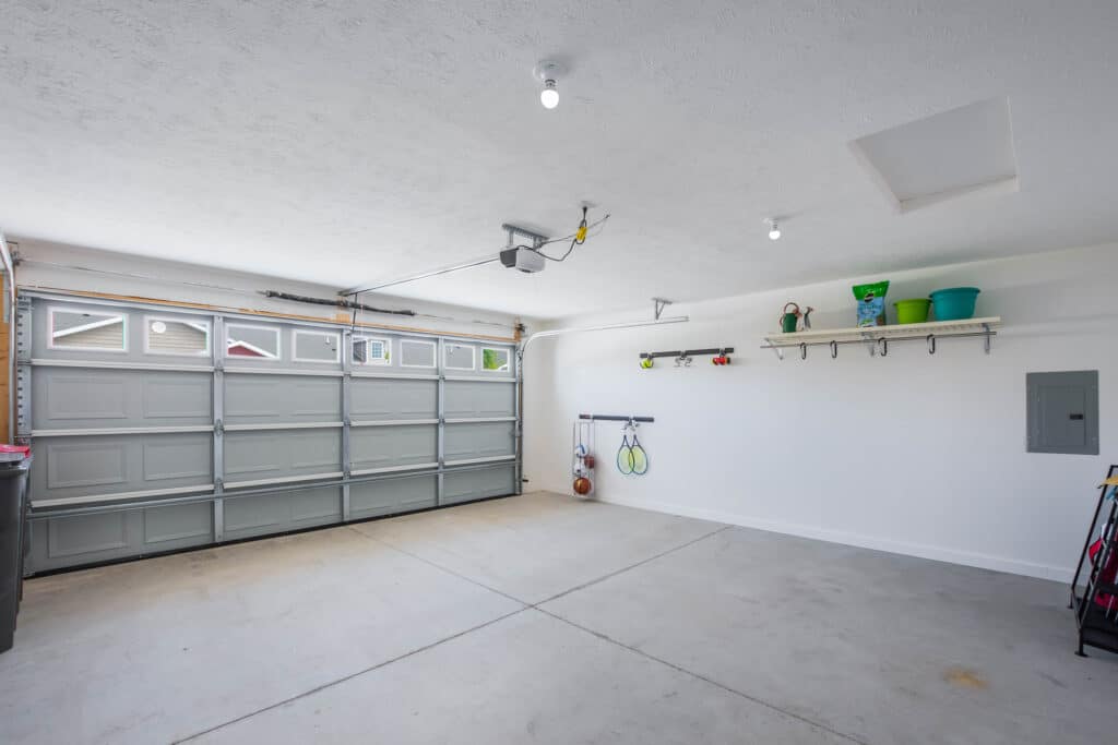 A clean, empty garage with a closed double garage door, wall-mounted shelves holding various items, and a few hanging tools and hose. The floor and walls are white, and the space is well-lit.