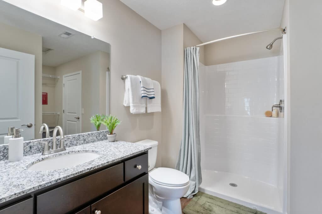 A bathroom with a granite countertop, sink, toilet, and a walk-in shower with a white tile wall and curtain. Towels hang on the wall, and a small plant is on the counter.