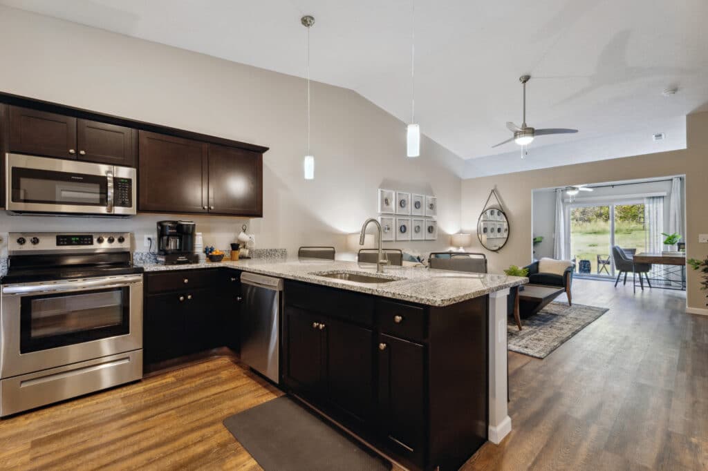 Modern kitchen with dark cabinets, stainless steel appliances, and a granite island. Open layout flows into a living area with a ceiling fan, wall art, and a dining table by glass patio doors.