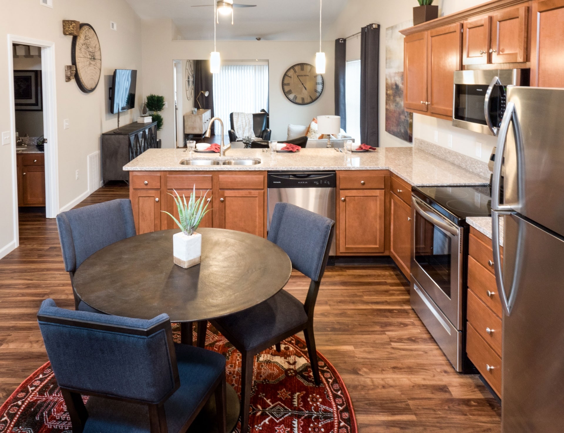 A modern kitchen with wooden cabinets, stainless steel appliances, and a central dining area. A round table with blue chairs is on a patterned rug. A large clock decorates the wall.