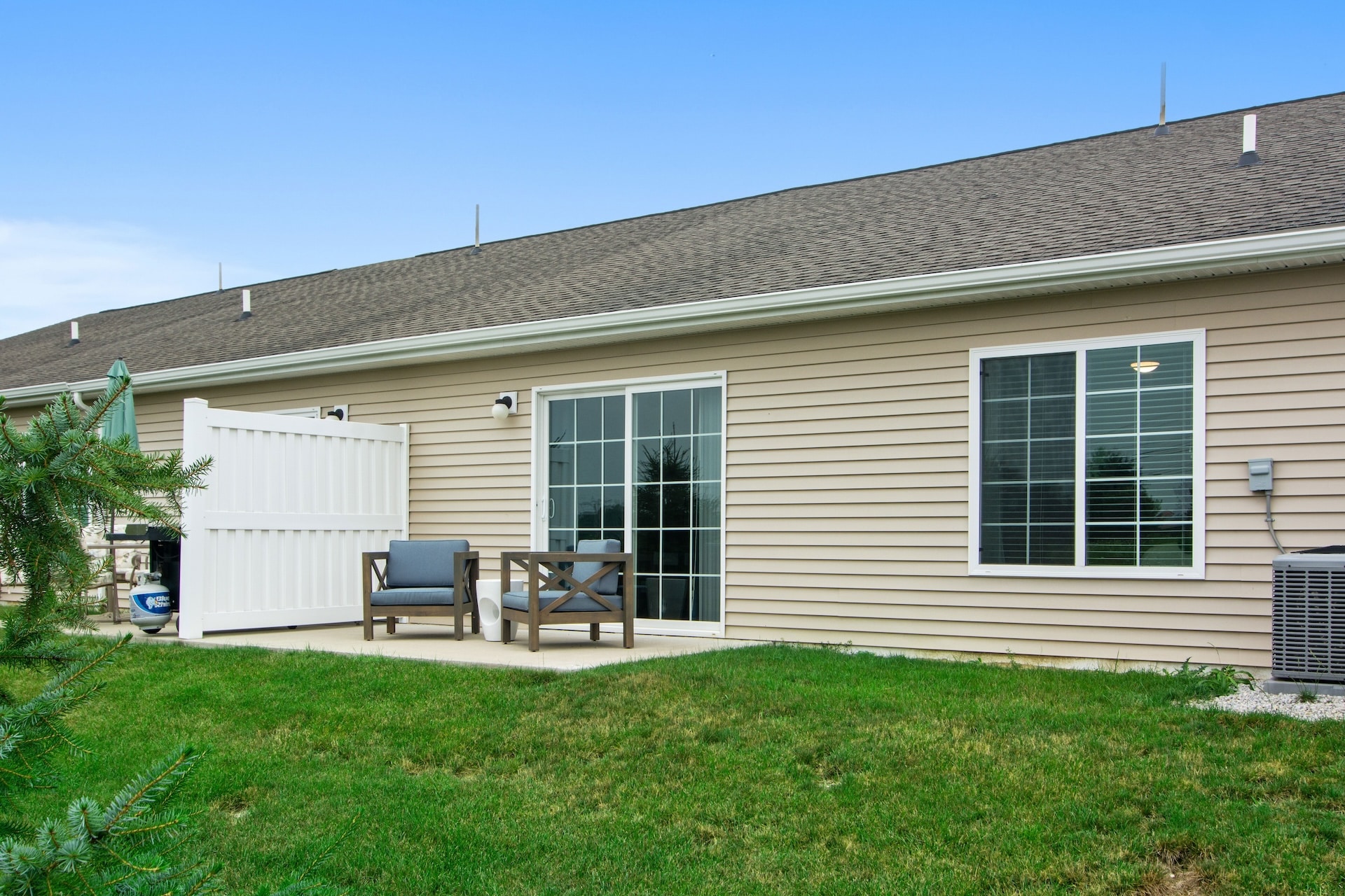 Patio area behind a beige siding building with a glass sliding door, two chairs, a table, and a small lawn. An air conditioning unit is visible on the right.