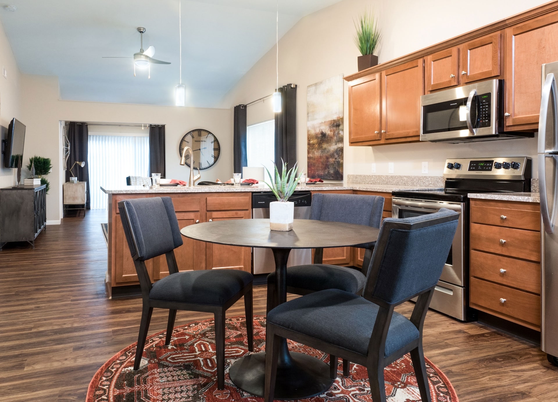 Open-concept kitchen and dining area with a round table, four chairs, wooden cabinets, stainless steel appliances, and a ceiling fan. A rug is under the table, and decor items are present.