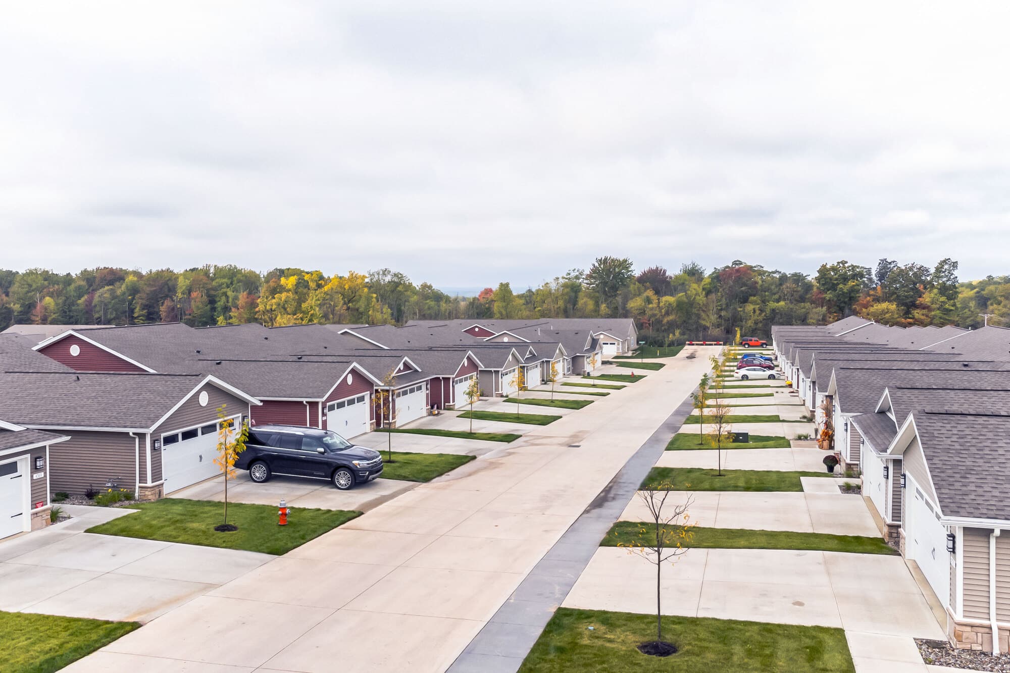 A row of single-story houses with garages, lined along a paved street, surrounded by green lawns and trees in the background.