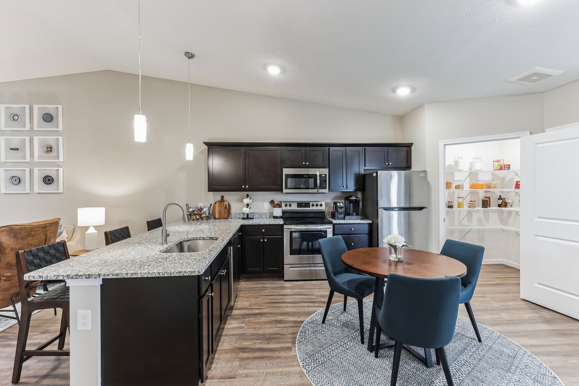 Modern kitchen with dark wooden cabinets, stainless steel appliances, granite countertops, and a round dining table with four chairs. A pantry with open shelving is visible in the background.