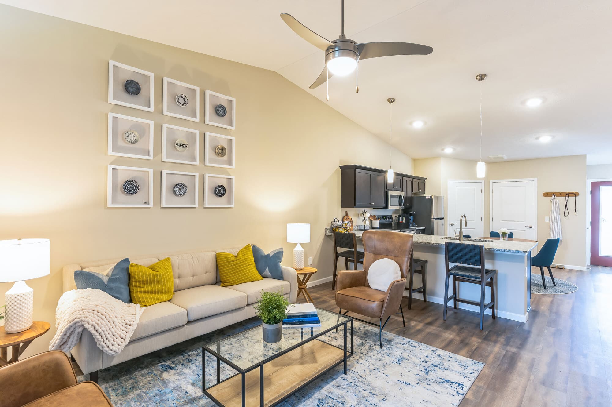 A modern open-concept living room and kitchen with a beige sofa, blue rug, and bar stools at the kitchen island. Decor includes framed wall art and pendant lights.