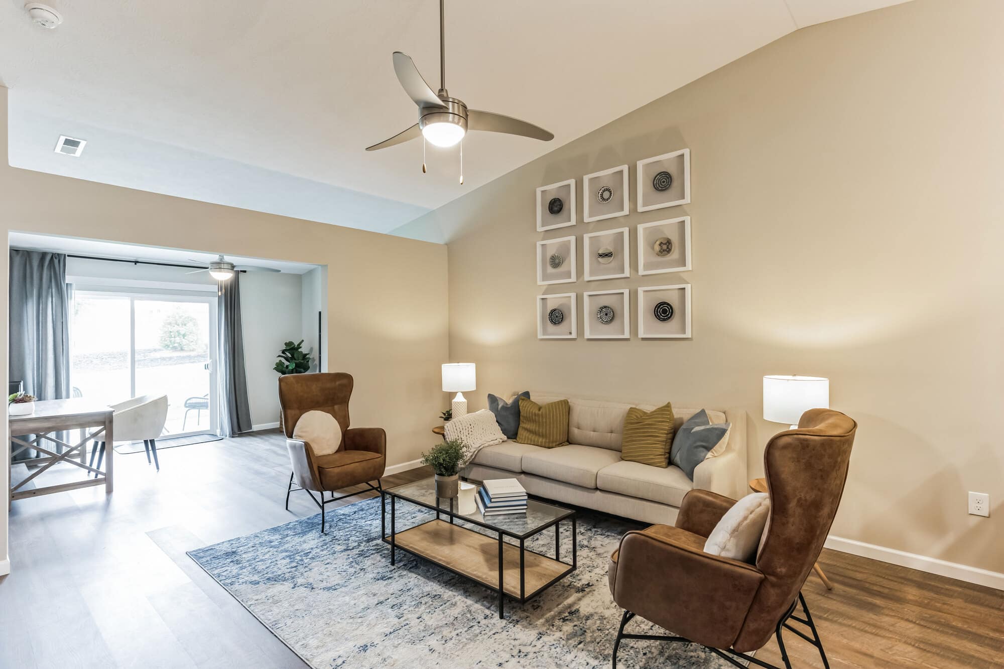 Modern living room with a beige sofa, brown armchairs, a glass coffee table, and wall art. Ceiling fan and lamps provide lighting. Dining area visible in the background.