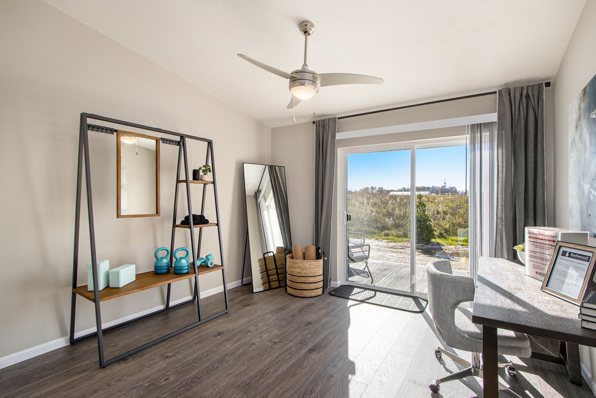 A bright, modern home office with a desk, sliding glass doors, exercise area with kettlebells, a full-length mirror, and wall-mounted shelving.