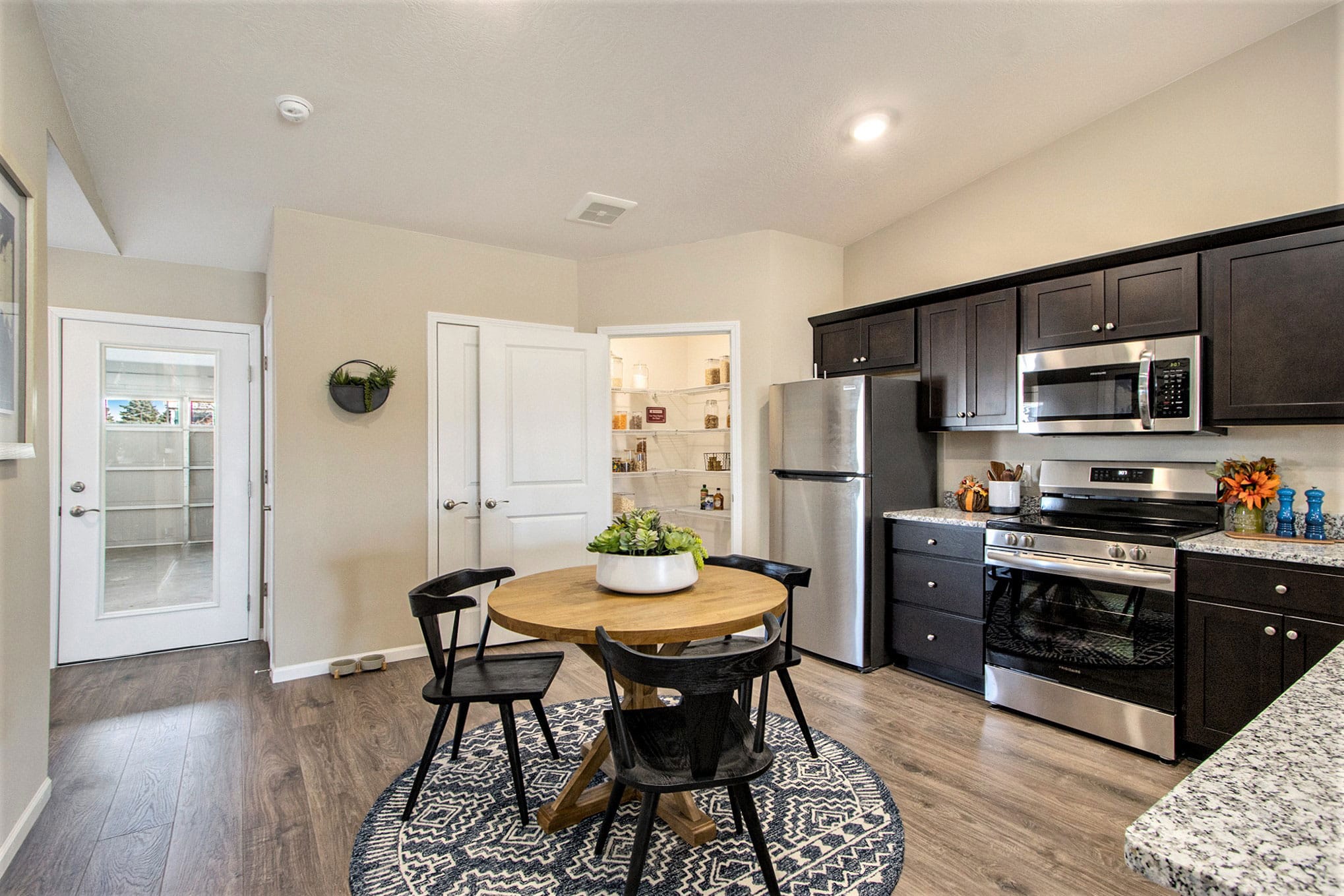 A modern kitchen with dark cabinets and stainless steel appliances. A round wooden table with four black chairs sits on a patterned rug. A plant decorates the table.