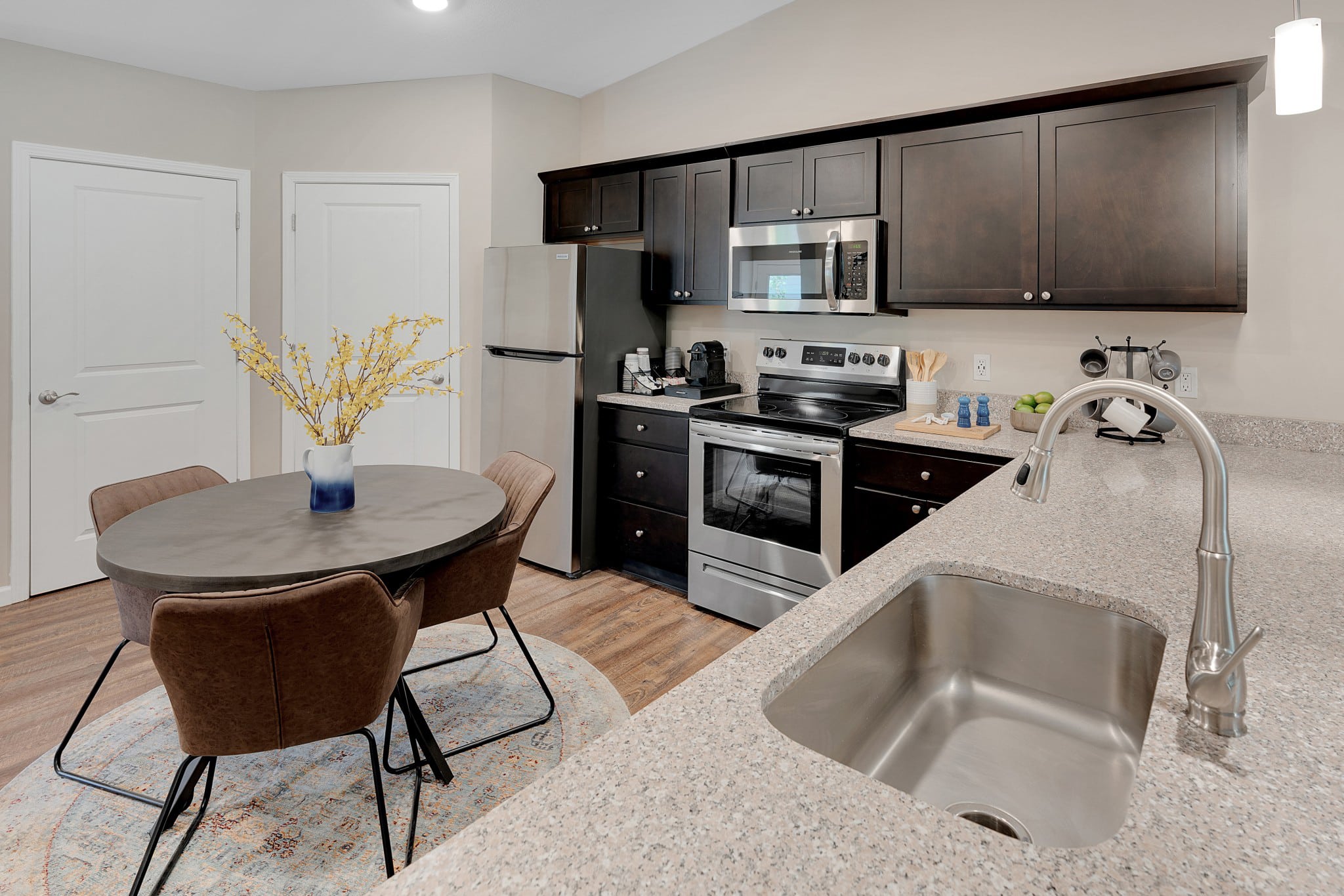 A modern kitchen with stainless steel appliances, dark cabinetry, a round dining table with chairs, and a sink in the foreground. An arrangement of yellow flowers is on the table.