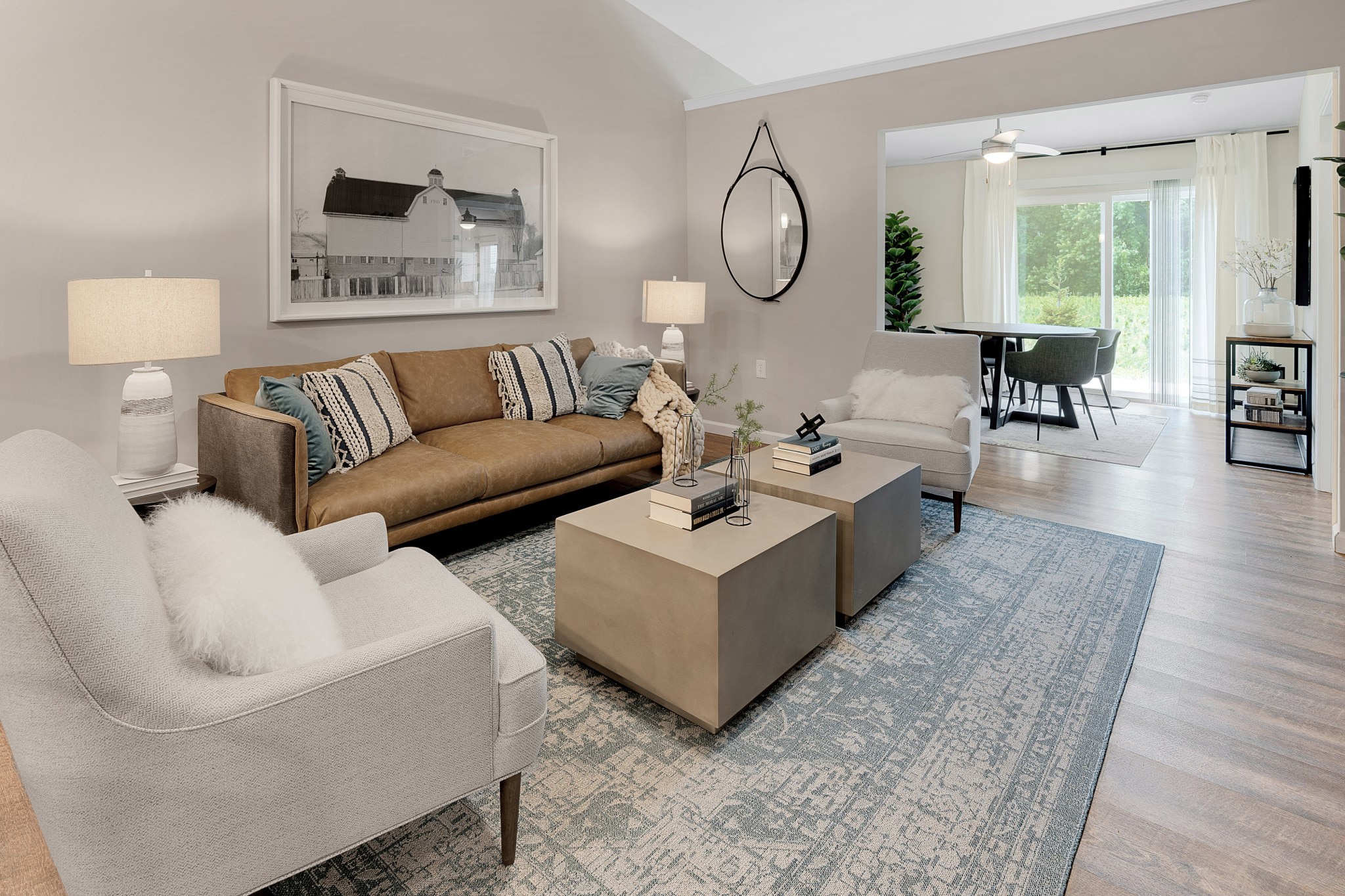 A modern living room features a brown sofa, two beige chairs, two square coffee tables, a large rug, wall art, table lamps, and a circular mirror on the wall. A dining area is visible in the background.