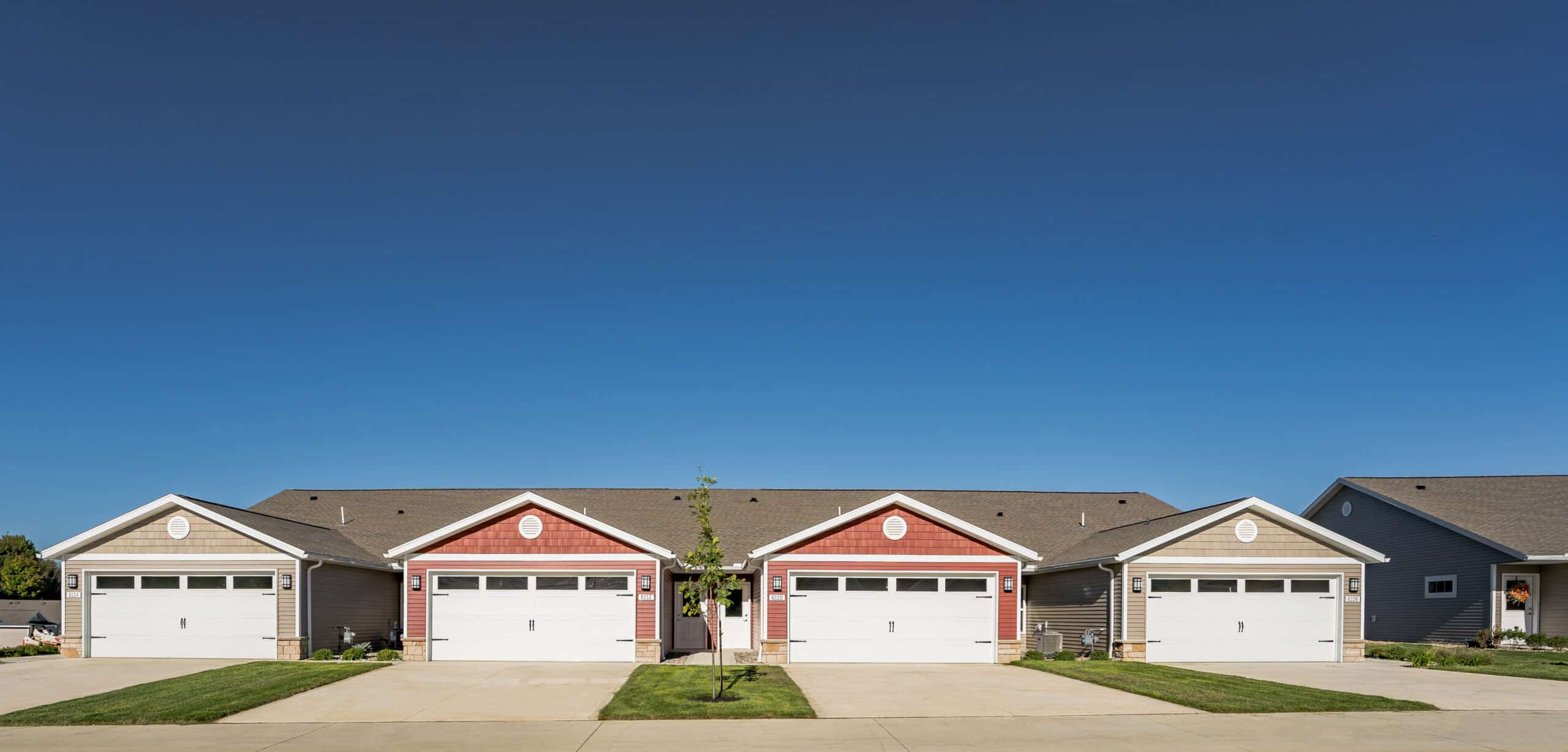 A row of single-story, connected townhomes with garages, in red and gray, under a clear blue sky, with well-kept lawns and a driveway in front.