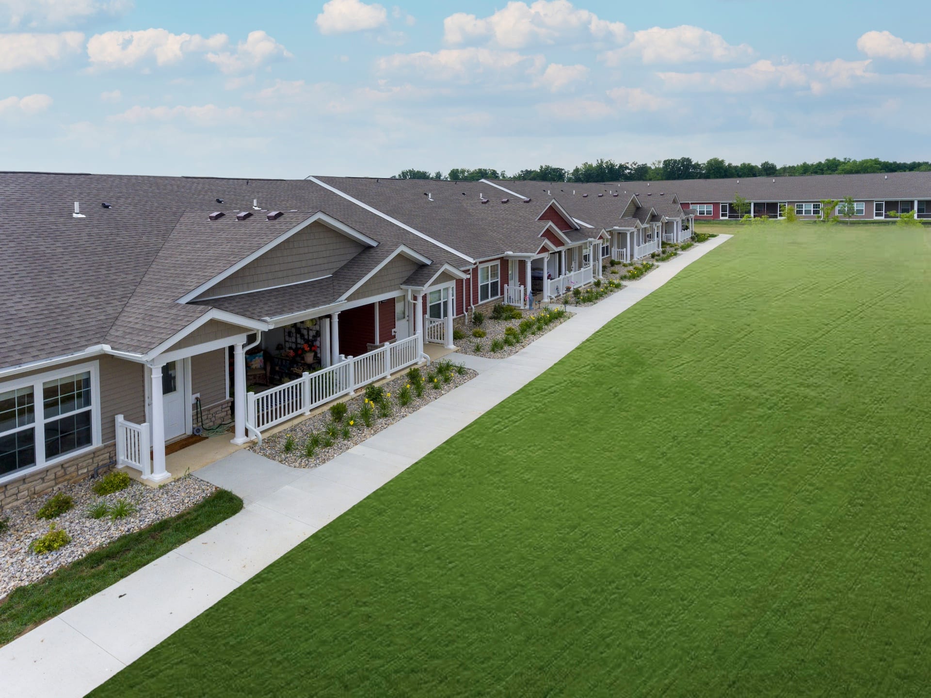 A row of single-story homes with front porches and lawns on a clear day. A large grassy area stretches out beside the homes.