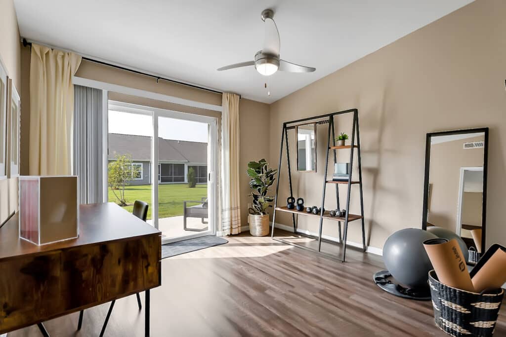 A bright room with a wooden desk, indoor plants, gym equipment, large standing mirrors, and a view of a garden through sliding glass doors.