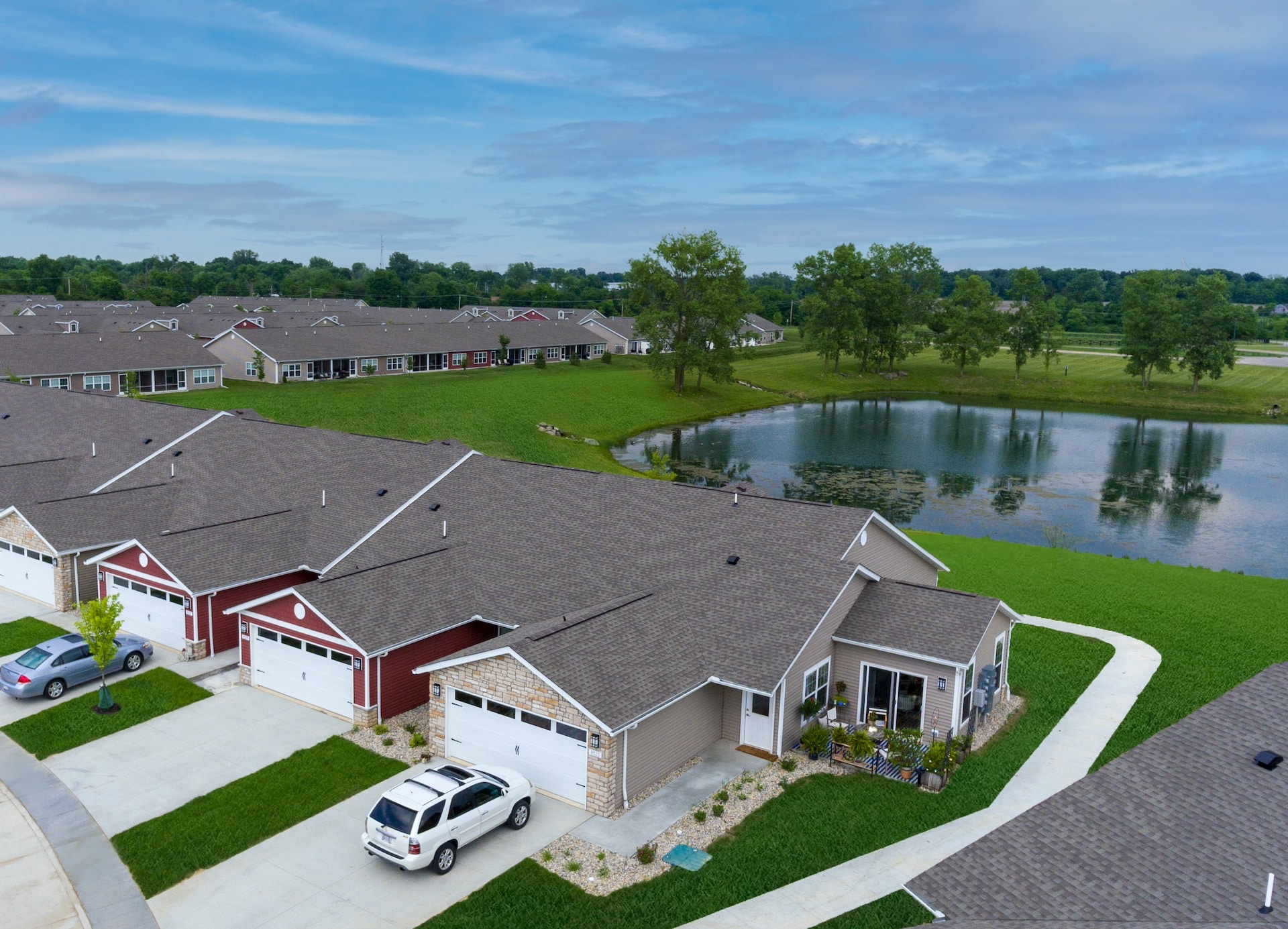 Aerial view of a suburban neighborhood with rows of single-story houses, some red and some beige, a white SUV parked in a driveway, and a pond with greenery in the background.