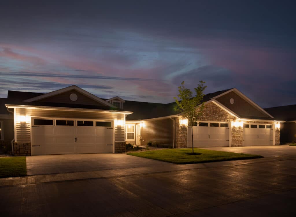 Two modern suburban houses with illuminated garages and driveways at dusk, featuring landscaped lawns and a tree in the foreground.