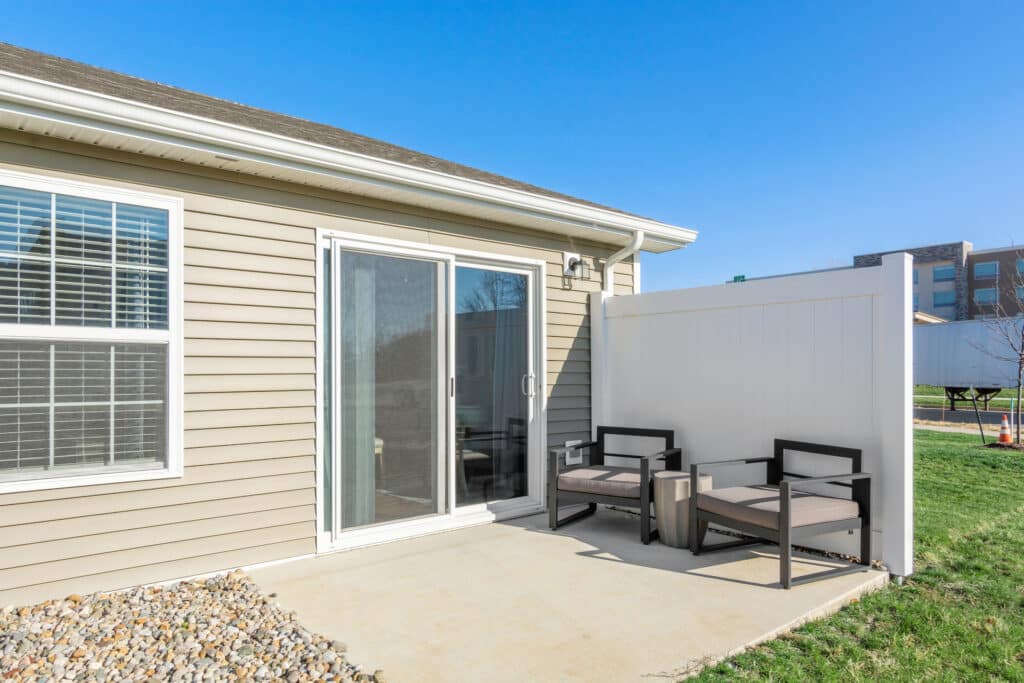 A small concrete patio with two cushioned chairs and a table beside a beige house with sliding glass doors. The area is enclosed by a white privacy fence. There is a rock bed along the house wall.