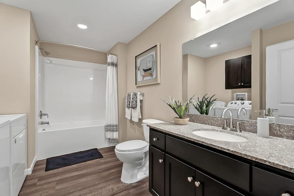 A modern bathroom with beige walls featuring a bathtub with a white shower curtain, a toilet, a double-sink vanity, a potted plant, and a mirror above the sink. A washing machine is visible in the background.