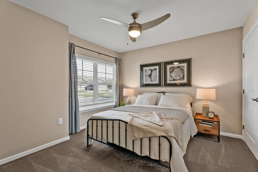 Bedroom with a large window, metal bed frame, white bedding, two bedside tables with lamps, beige walls, ceiling fan, framed artwork above the bed, and a carpeted floor.