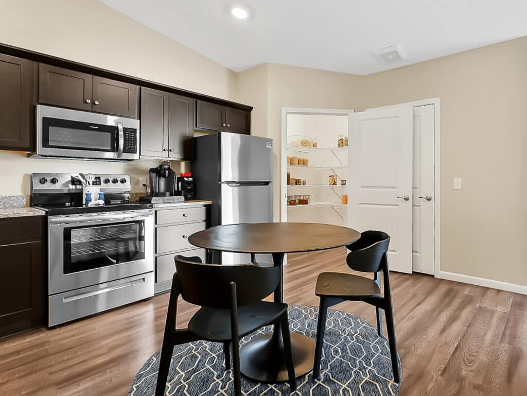 Modern kitchen with dark cabinets, stainless steel appliances, round dining table with three black chairs, and a pantry with open double doors and visible shelves filled with items.