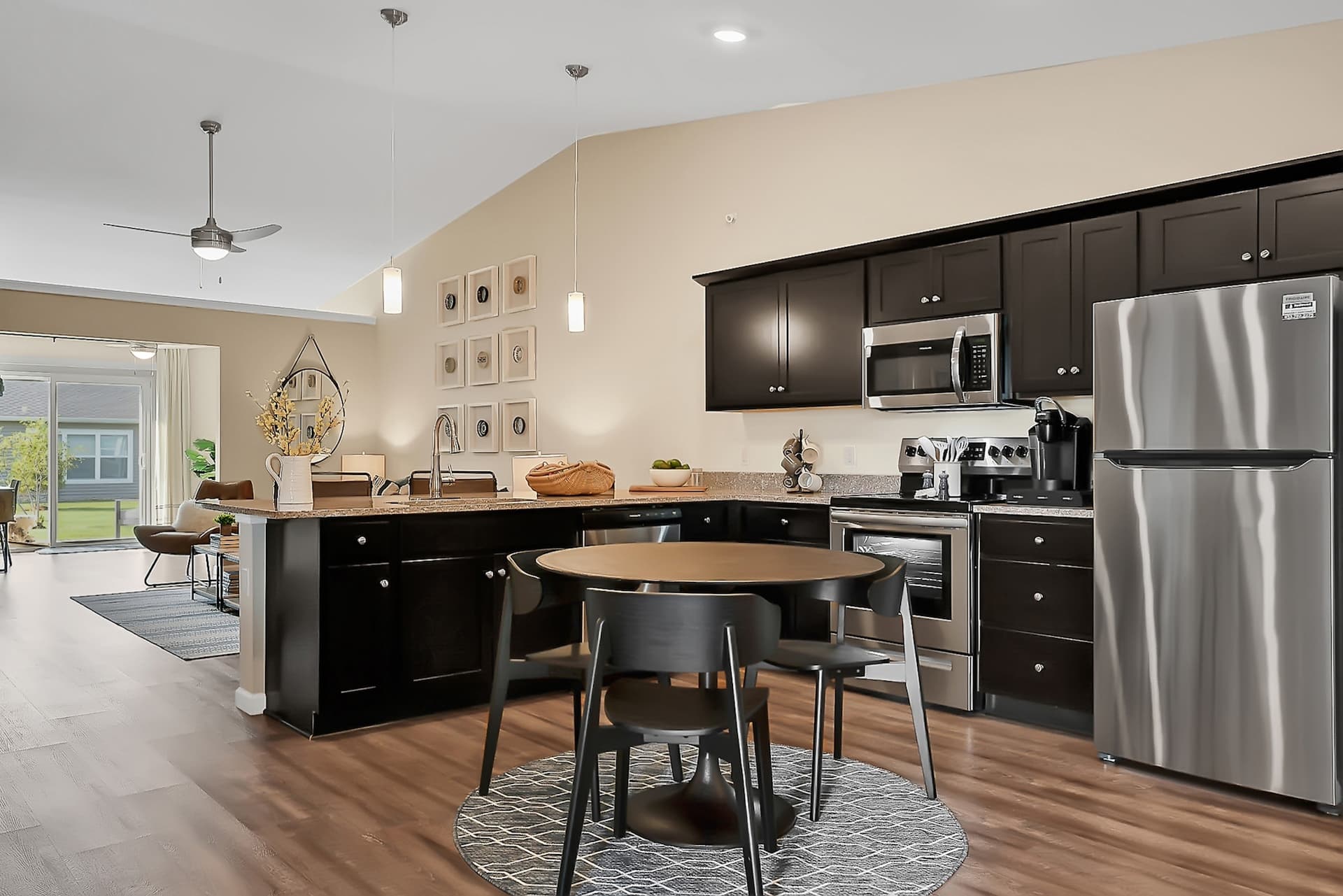 Modern kitchen with dark wood cabinets, stainless steel appliances, and a round dining table with chairs on a patterned rug. Open layout extends to living area with natural light through large windows.