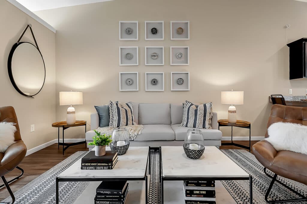 A modern living room with a gray sofa, two armchairs, a round wall mirror, framed artwork, table lamps, and two coffee tables decorated with books and plants.