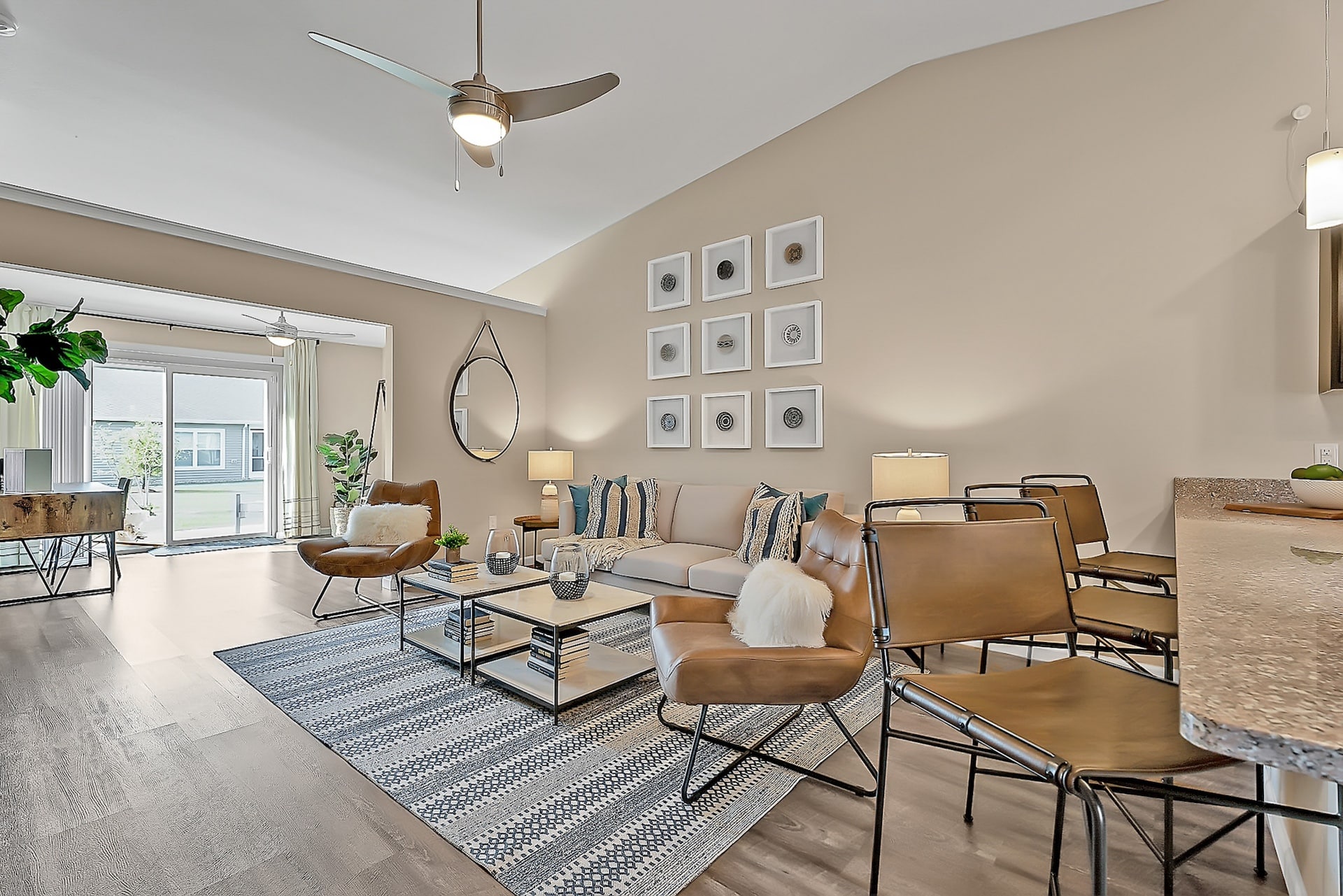 Modern living room with neutral tones featuring a beige sofa with pillows, brown leather chairs, a striped rug, wall art, glass coffee table, bar stools, plants, and large windows with natural light.