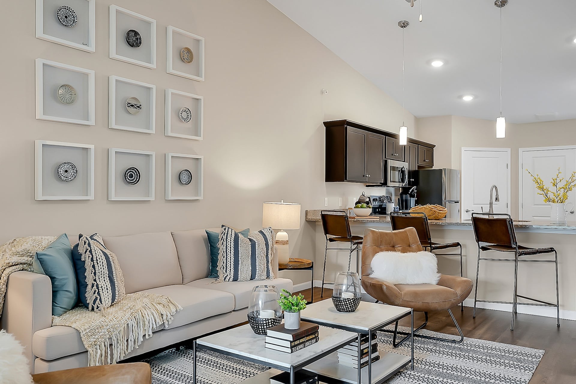 Modern living room with a beige sofa, decorative pillows, and framed wall art. A kitchen with dark cabinets, pendant lights, and a breakfast bar with stools is visible in the background.