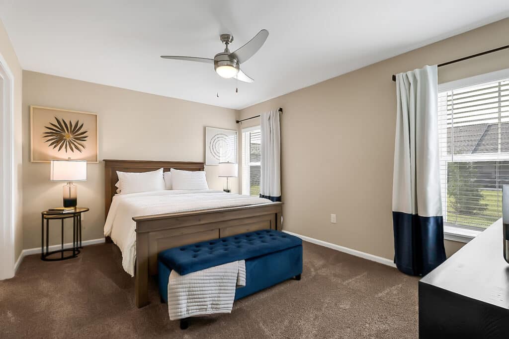 A bedroom with beige walls features a wooden bed with white bedding, a blue bench at the foot, a ceiling fan, two windows with curtains, and a nightstand with a lamp.