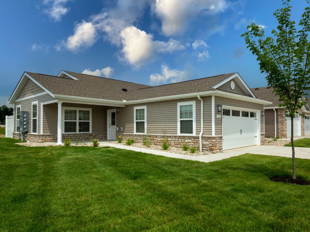 Single-story house with beige siding, white trim, and a double garage. The front yard has a manicured lawn, young tree, and small flower beds. The sky is partly cloudy.