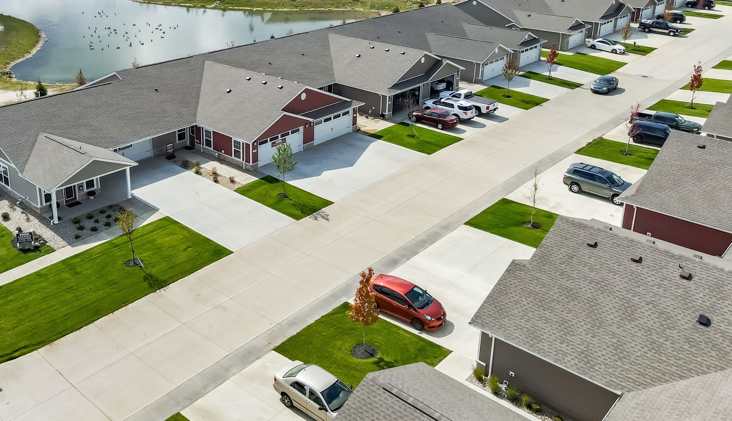 Aerial view of a suburban neighborhood with identical houses, driveways, and parked cars. A small pond is visible in the background.