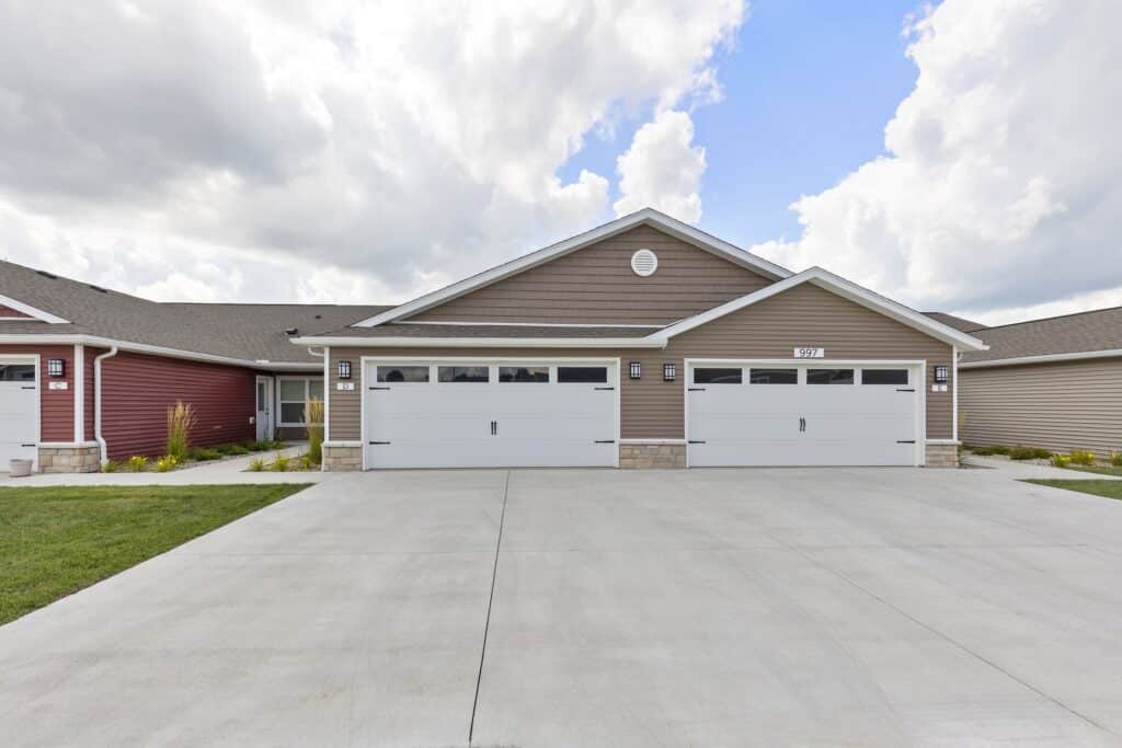 Duplex building with two separate garages, beige siding, white trim, and a wide concrete driveway under a partly cloudy sky.