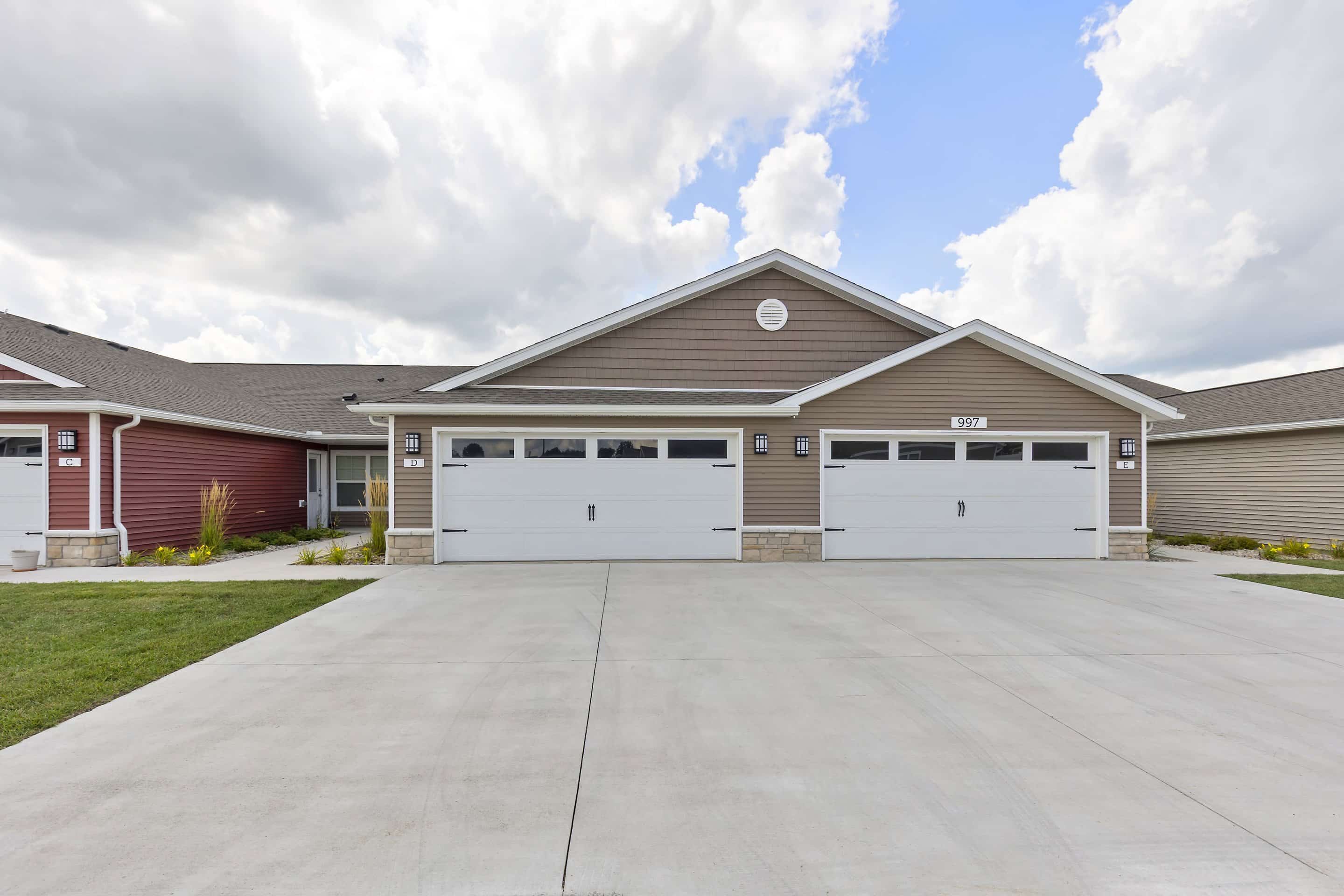Duplex building with two separate garages, beige siding, white trim, and a wide concrete driveway under a partly cloudy sky.