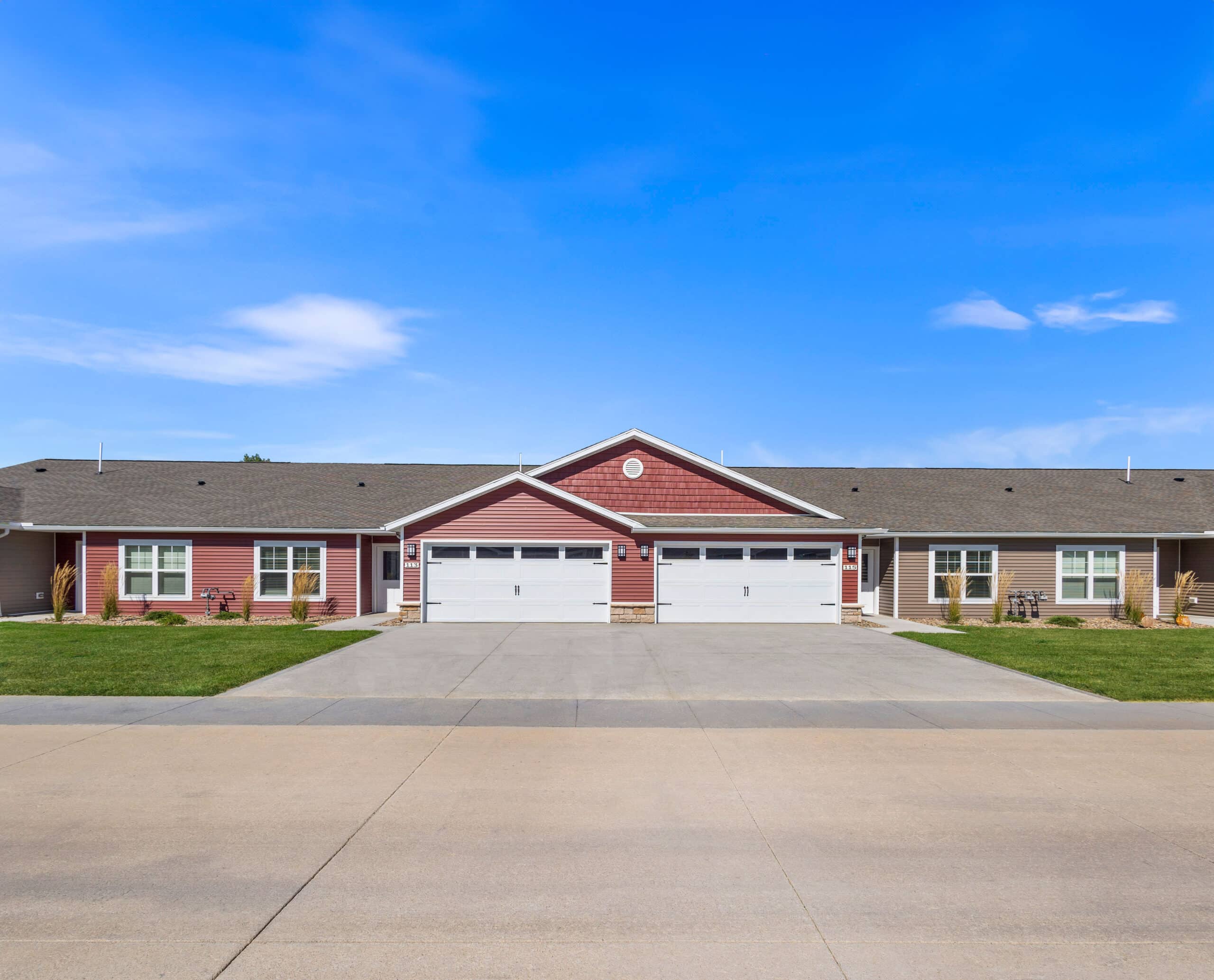 A duplex with red and gray siding, featuring two garage doors and a wide driveway, set against a clear blue sky.