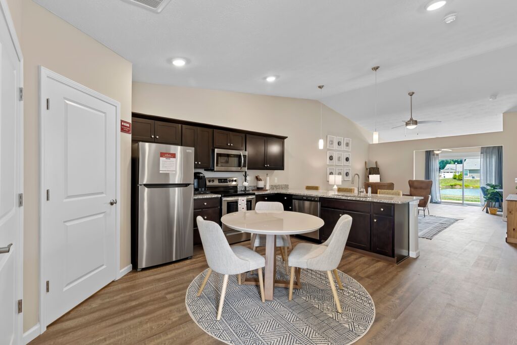 Modern kitchen and dining area with dark cabinets, stainless steel appliances, round dining table, four chairs, and an open view into a living room with large windows.