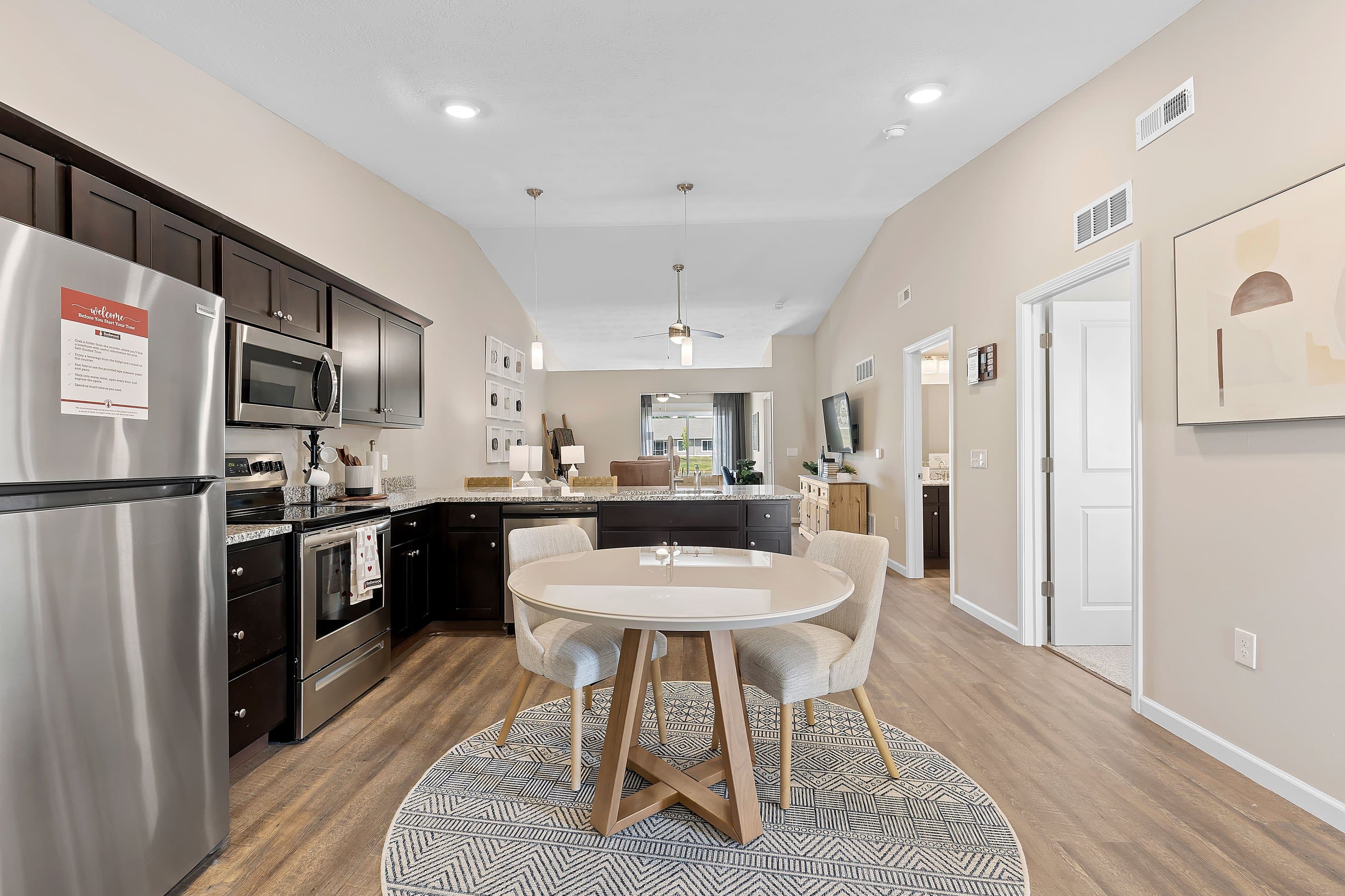 Modern kitchen and dining area with stainless steel appliances, dark wood cabinets, round table with two chairs on a rug, and open view into living room.