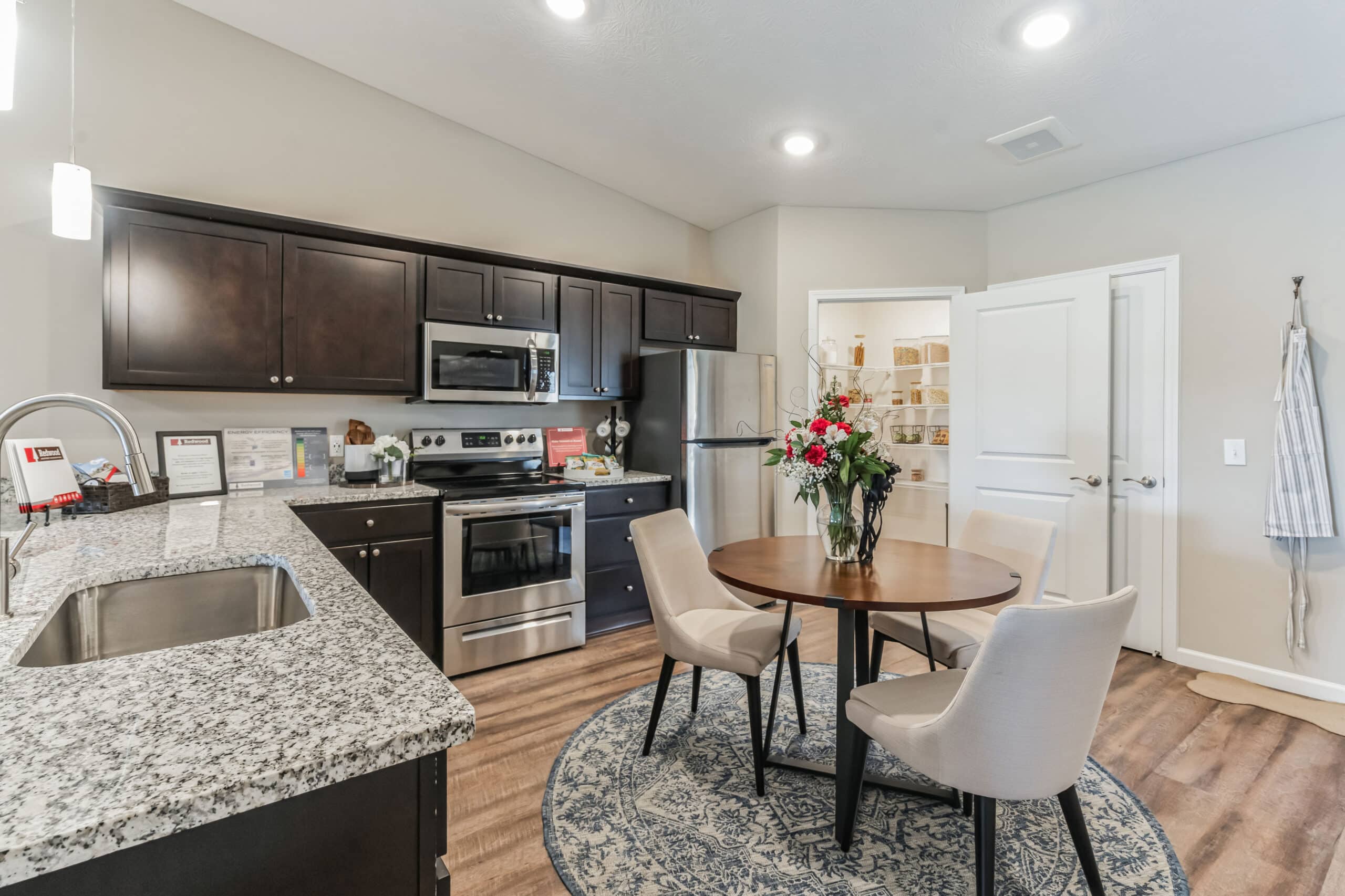 Modern kitchen with dark cabinets, granite countertops, stainless steel appliances, and a round dining table with four chairs. A vase with flowers sits on the table.