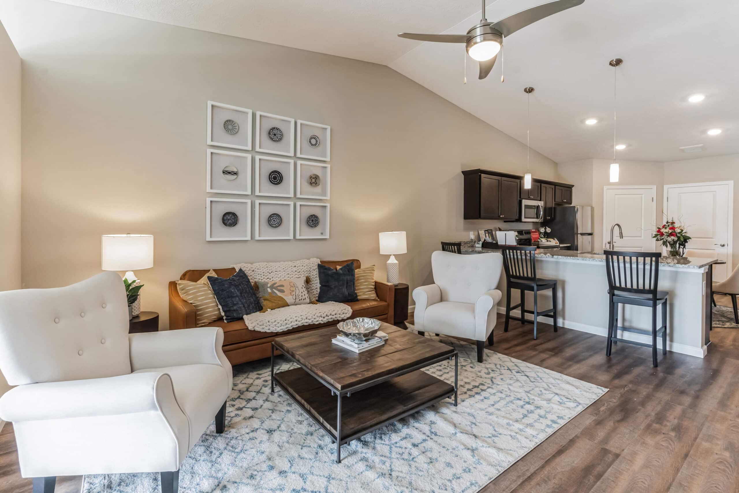 A modern living room and kitchen with a brown sofa, white armchairs, a wooden coffee table, and a kitchen island with bar stools. Neutral tones and decorative wall art are present.